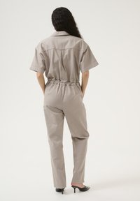 Ljust grå jumpsuit med korta ärmar, elastisk midja och smalnande ben. Slen tyg och minimalistisk design. Bärs med svarta klackar.