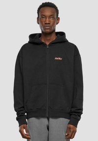 Crna hoodie s patentnim zatvaračem od mješavine pamuka. Ima prednje džepove, rebraste manžete i kapuljaču. Mali crveni logotip na lijevoj strani prsnog koša.
