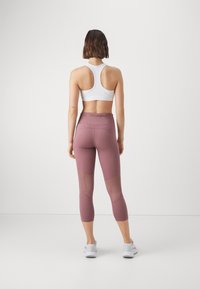 Vrouw die met de rug naar de camera staat, gekleed in een witte sportbeha, mauve cropped legging met meshpanelen en witte sport schoenen, op een witte achtergrond.