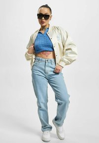 Lys kremfarget sateng bomberjakke, blå strikket crop top, lyseblå vid jeans og hvite sneakers. Merkelig tatovering på midjen. Sorte solbriller.