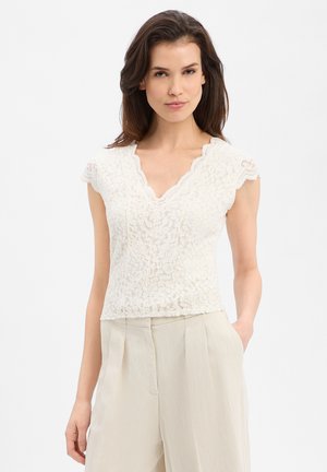 Frau trägt ein weißes Spitzen-Top mit kurzen Ärmeln und geschwungenem Ausschnitt sowie beige, hoch Taillen-faltenhose, steht mit einer Hand in der Tasche.