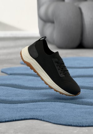 Chaussure de sport noire avec tige en maille, accents texturés, design à lacets, semelle rembourrée et semelle extérieure en caoutchouc gomme, posée au-dessus d'un tapis bleu.