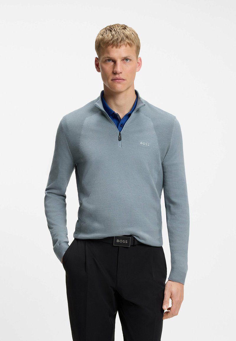 Homme debout les mains dans les poches, portant un pull bleu clair à fermeture quart de zip sur une chemise bleue, un pantalon noir et une ceinture noire BOSS.