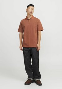 Rudas polo marškinėliai su apykakle, derinami su tamsaus denim medžiagos džinsais ir rudais batais. Marškiniai turi trumpas rankoves ir krūtinėje esantį logotipą.