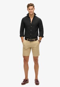 Camisa negra de lino con cuello doblado, pantalones cortos beige, cinturón marrón y zapatos burdeos. Diseño sencillo sin patrones visibles.