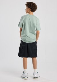 Light green cotton t-shirt with a relaxed fit, paired with knee-length black cargo shorts featuring two side pockets, worn with white sneakers.