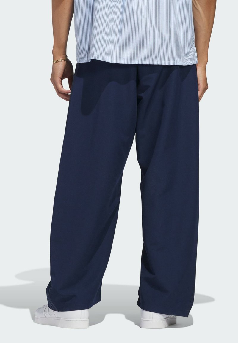 Persona que lleva pantalones holgados azul marino, zapatillas blancas y una camiseta azul clara a rayas, vista desde la cintura hacia abajo, de espaldas.