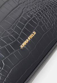 Zwarte tas van kunstleer met krokodillenstructuur, voorzien van een gouden "ANNA FIELD" logo. Soepele textuur met duidelijke schubben en randen.