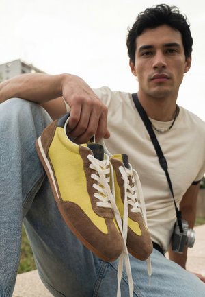 Junger Mann hält gelbe und braune Sneakers mit weißen Schnürsenkeln, trägt ein weißes T-Shirt und Jeans, sitzt im Freien mit einem Kameragurt über der Schulter.