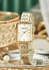 Reloj cuadrado de metal dorado y plateado con números romanos, fondo crema y pulsera texturizada. Presenta el nombre de la marca "Victoria-Hyde" y detalles intrincados.
