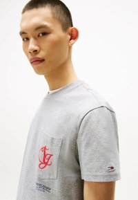 Camiseta de algodón gris con un bolsillo tipo parche que presenta un logo rojo. Manga corta, cuello redondo y un sutil acento de la marca en la manga.