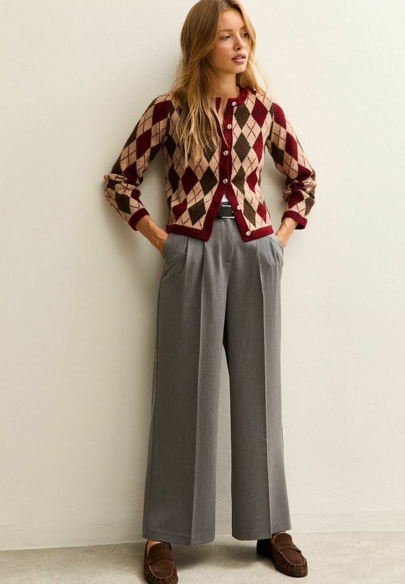 Cardigan a rombi rosso e beige con bottoni, abbinato a pantaloni a gamba larga grigio chiaro e mocassini marroni. Sfondo semplice.