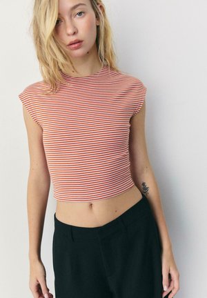 Donna bionda che indossa un top corto a righe rosse e bianche e pantaloni neri, in piedi davanti a uno sfondo chiaro e uniforme.