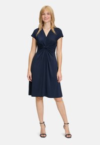 Robe bleu marine mi-longue avec un décolleté en V, des manches courtes, et un détail de nœud tordu à la taille, fabriquée en tissu doux et extensible.