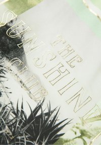 Textil con "EL CLUB DEL SOL" en relieve dorado sobre un fondo gráfico verde y negro, con patrones de hojas negras afiladas en la parte inferior.