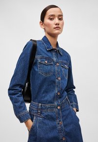 Mørkeblå denimskjortedress med krage, lange ermer, to brystlommer og knappestolpe. Inkluderer broderte detaljer og markert midje.