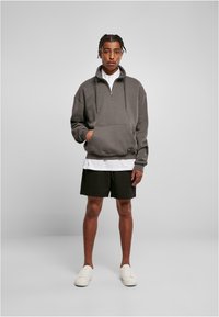 Grå hettegenser med glidelås og frontlomme, båret over en hvit skjorte og svarte shorts. Har ribbestrikkede mansjetter og en avslappet passform. Hvite joggesko.