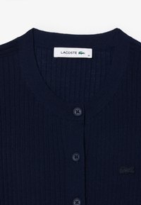 Cardigan a coste navy con scollo rotondo, sei bottoni scuri e logo Ricamato Lacoste sul lato sinistro del petto. Taglia: 36.