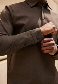 Cămașă lungă, cu mânecă lungă, de culoare maro, dintr-un material texturat, cu guler standard și manșete cu butoane. Prezintă cusături subtile și câte două butoane pe fiecare manșetă.