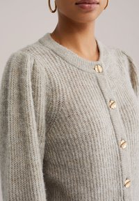 Světlý šedý pletený cardigan s žebrovanou texturou, kulatým výstřihem a pěti knoflíky ve zlaté barvě. Tkanina vypadá měkce a teple.