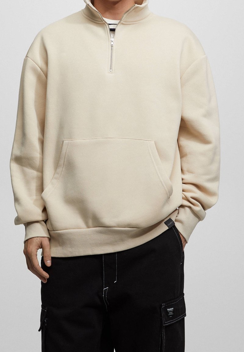 Beige quarter-zip pullover med en frontlomme, ribbede ærmeafslutninger og en blød tekstur. Kombineret med sorte cargobukser med sidelommer.