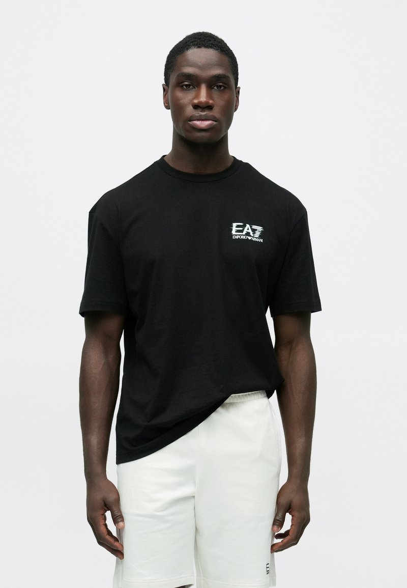 Férfi modell fekete EA7 Emporio Armani pólót és fehér rövidnadrágot visel, egyszerű, világos háttér előtt áll, előre nézve.