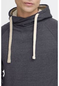 Sweat à capuche en tissu gris foncé avec une finition texturée ; doté de cordons de serrage crème et d'un subtil accent triangulaire au niveau de l'encolure.