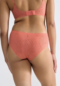 De rug van een vrouw die een bijpassende koraalroze beha en hoge taille slipjes draagt met een subtiel gestructureerd patroon op een neutrale achtergrond.
