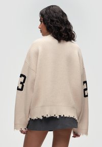 Beiger oversized Pullover mit geripptem Textur, featuring einen fransigen Saum und einer schwarzen Nummer 23 auf dem Ärmel. Das Modell trägt einen dunklen Rock.