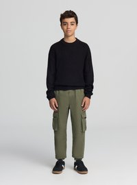 Czarny dziergany sweter, oliwkowozielone spodnie cargo z kieszeniami oraz czarne trampki. Strój charakteryzuje się luźnym krojem i fakturowaną tkaniną.