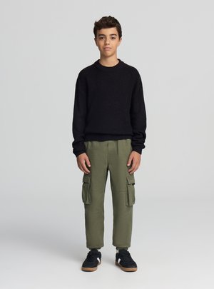 Maglione lavorato a maglia nero, pantaloni cargo verde oliva con tasche e sneakers nere. L'outfit presenta una vestibilità ampia e un tessuto testurizzato.