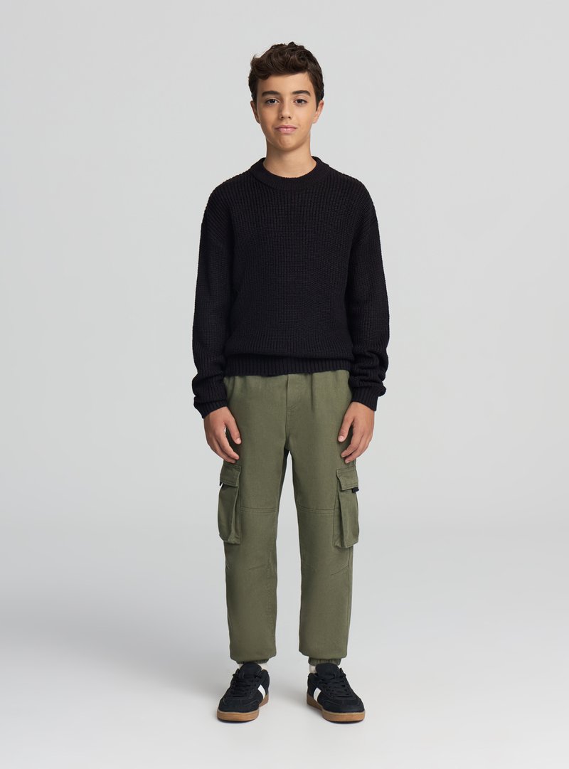 Czarny dziergany sweter, oliwkowozielone spodnie cargo z kieszeniami oraz czarne trampki. Strój charakteryzuje się luźnym krojem i fakturowaną tkaniną.