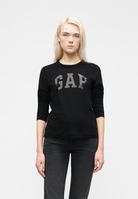 Ξανθιά γυναίκα που φοράει μαύρο μακρυμάνικο μπλουζάκι GAP και σκούρο τζιν, που στέκεται μπροστά σε ένα απλό λευκό φόντο.