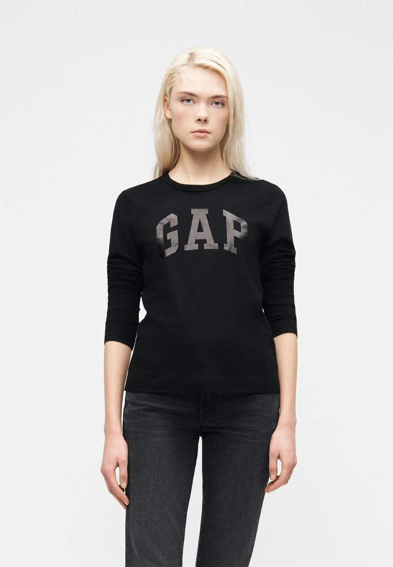 Ξανθιά γυναίκα που φοράει μαύρο μακρυμάνικο μπλουζάκι GAP και σκούρο τζιν, που στέκεται μπροστά σε ένα απλό λευκό φόντο.