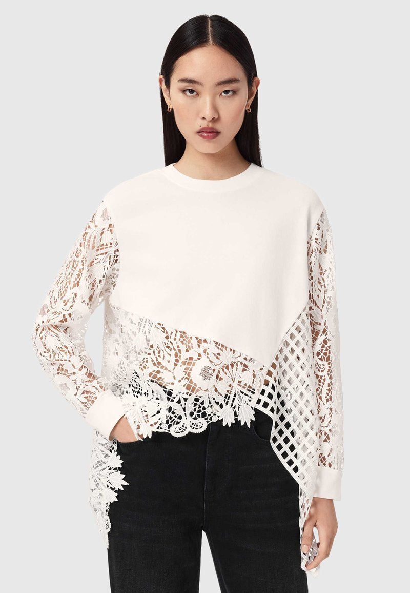 Jeune femme aux longs cheveux foncés portant un haut blanc avec des panneaux mixtes unis et en dentelle florale, associé à un pantalon noir, en position neutre.