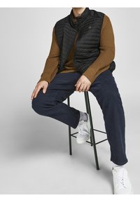 Czarna pikowana kamizelka na brązowym swetrze o fakturze, w połączeniu z ciemnoniebieskimi spodniami i białymi trampkami, siedząca na zielonym metalowym stołku.