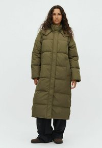 Woman with long curly hair wearing a long olive green puffer coat, black pants, and brown shoes, standing against a plain light background.