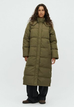 Woman with long curly hair wearing a long olive green puffer coat, black pants, and brown shoes, standing against a plain light background.