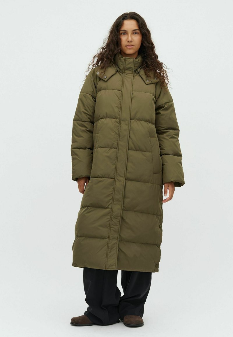 Woman with long curly hair wearing a long olive green puffer coat, black pants, and brown shoes, standing against a plain light background.