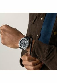 Sølv ur med struktureret sort skive og chronograph-funktioner, silikonerem, båret på håndleddet sammen med et sort perlearmbånd.