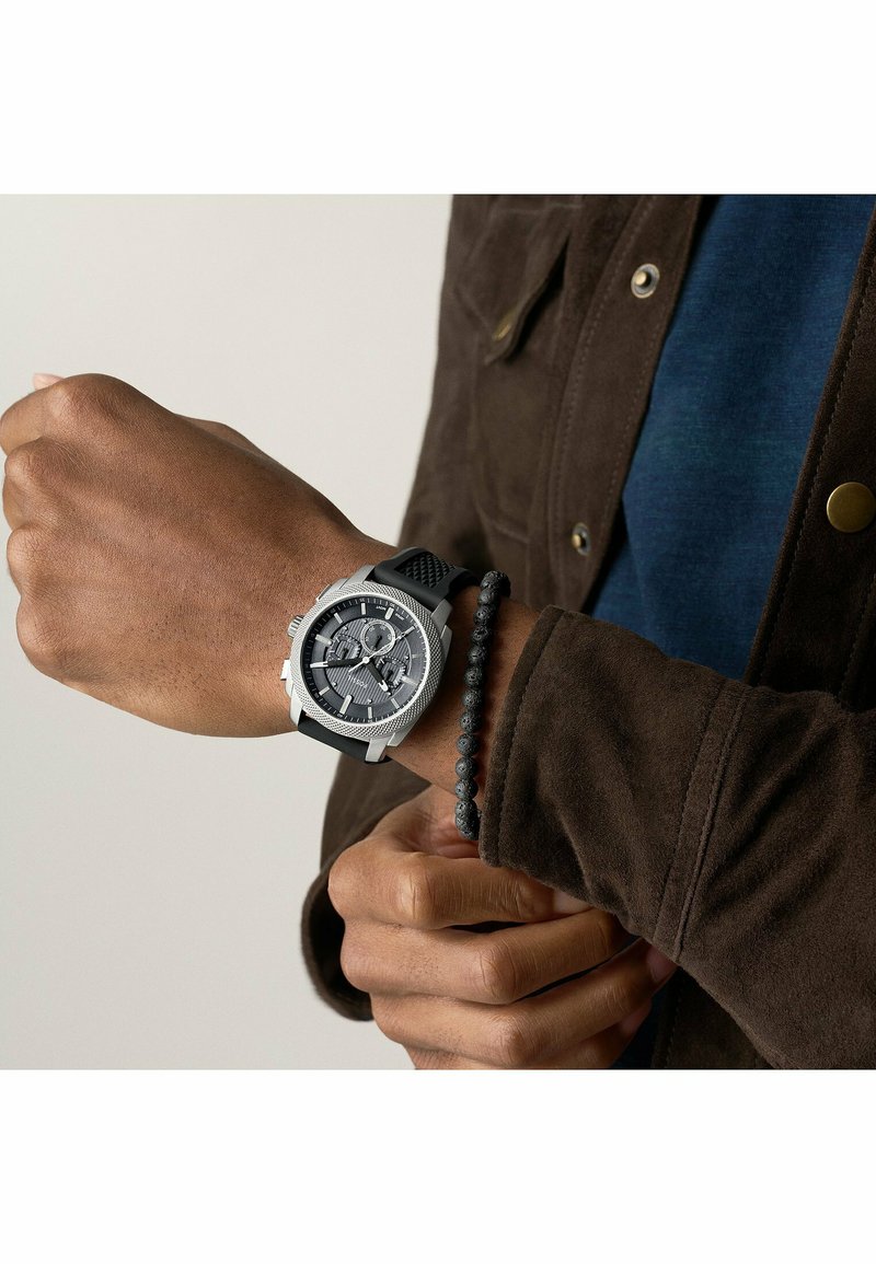 Sølv ur med struktureret sort skive og chronograph-funktioner, silikonerem, båret på håndleddet sammen med et sort perlearmbånd.