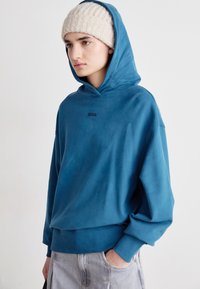 Felpa con cappuccio blu dal morbido tessuto, tasca frontale e polsini a coste. Logo "BOSS" nero centrato sul petto. Indossata con pantaloni shorts in denim grigio chiaro.