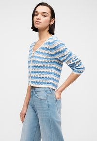 Νεαρή γυναίκα με κοντά σκούρα μαλλιά φοράει ένα πλεκτό ζακέτα με μπλε και άσπρες ρίγες και ανοιχτόχρωμα τζιν ψηλής μέσης, με το χέρι στην τσέπη.