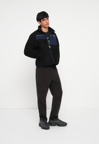 Schwarze Fleecejacke mit navyblauen Akzenten, mit Reißverschluss und hohem Kragen. Kombiniert mit dunklen Cordhosen und schwarzen Turnschuhen.
