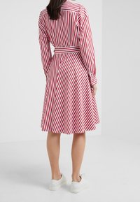 Czerwona i biała sukienka koszulowa w paski, z długimi rękawami, talią związaną w pasie i spódnicą w kształcie litery A, zestawiona z białymi trampkami. Gładka tekstura materiału.