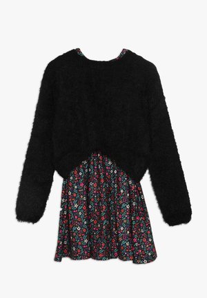 Sort, fnugget trøje med lange ærmer lagdelt over en kort blomsterkjole med små blomster i flere farver på en mørk baggrund.