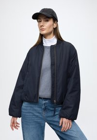 Tamsiai mėlynas bombonešio stiliaus švarkas su užtrauktuku priekyje, ribyta apykakle ir tekstūruota medžiaga, derinamas su pilku džemperiu ir džinsais. Juoda kepurė papildo išvaizdą.