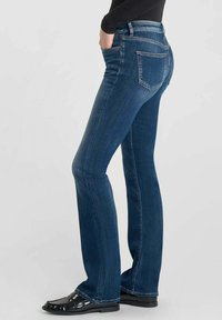 Persona vistiendo jeans azul de corte amplio y mocasines negros, con una mano en el bolsillo, mostrado de lado sobre un fondo simple.