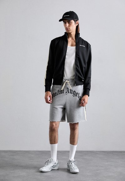 Young man wearing black zip-up jacket, gray shorts with "Palm Angels" text, white tank top, black cap, white socks, and sneakers standing on gray floor.