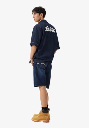 Uomo che indossa una camicia blu scuro a maniche corte con la scritta "Basic" sulla schiena, pantaloncini di jeans abbinati, calzini neri e stivali da lavoro color beige, in piedi di lato.